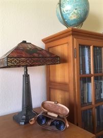 Glass front teak bookcase which matches the office desk and pair of filing cabinets. Modern Craftsman-style lamp with cowboy riding his horse motif. Antique pair of field glasses in leather case. Vintage world globe on stand. 