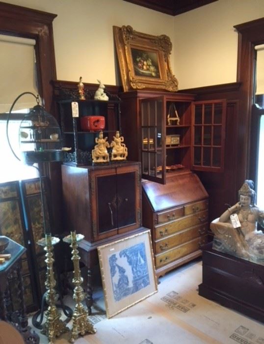 A view of the northwest corner of the dining room, showing massive antique brass candlesticks, a framed Thai or Cambodian print or rubbing, a pair of Chinese soapstone figures, a Japanese lacquerware tiered box, a vintage standing birdcage, a beautiful 2-part wood cabinet/desk ensemble, and so on. 