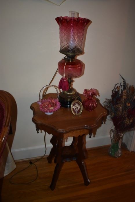 Gorgeous and Rare Antique Aladdin Style Cranberry Oil Lamp featuring Cranberry Bowl and Cranberry Fluted Edged Lamp Shade notice also the Cranberry Fluted Edged Vase and the Fenton Glass Basket with Cranberry and Milk Glass "Case Glass" with Handle. This all sets atop a Highly Carved Victorian Table with Scalloped Edging and Tear Drop Finials. Also is that a Cut Glass Pitcher I see on the floor to your right?