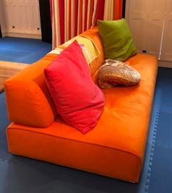 31) Pas Si Classique Armless Sofa from Ligne Roset Upholstered in Orange Ultrasuede. Two loose cushions, one in pink ultrasuede and one in acid green ultrasuede.