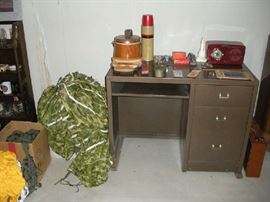 Basement:  A brown vintage metal desk displays a round canister shoe shine kit; shoe brushes; a thermos; some office supplies, and a vintage red BAKELITE radio.  