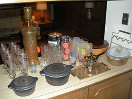 Kitchen:  Displayed are multiple vintage sets of LIBBY glass and mid-century glassware.  In the foreground are two pieces of matte black/gold PYREX.  To the right are two more pieces of PYREX that are gold/brown/fruit pattern AND a milk chocolate/gold/rooster pattern.  In the background is a tall amber color figural decanter.