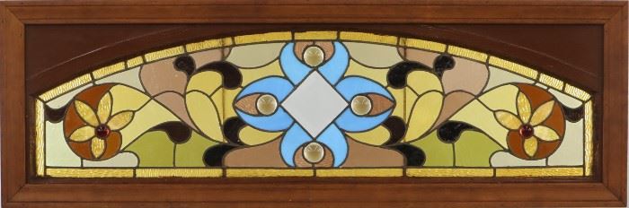 A late 19th century Stained Glass Transom Window.  Multi-color foliate design with cabochons and beveled glass accents in a wood frame.  Some cracked panels, minor frame damage.  57 1/2 x 19" high. 