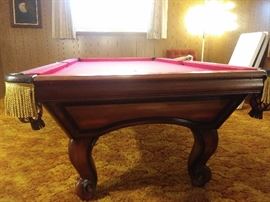 Custom Oak Pool Table by Golden West Billiards with accessories - the rectangular top having a plaque reading "Custom Made by Golden West Billiards, Inc. Los Angeles, CA", surrounding a red or maroon felt playing surface, the rail with leather and scarlet bullion fringe pockets, above shaped paneled sides, the whole rising on cabriole legs, ending in scroll feet. 32" high X 109" wide X 59" deep together with 1 cover, set of balls, cue sticks, racks & cue ball, 