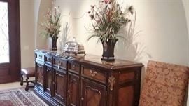 this extra long buffet used as a striking entry piece is by Marg Carson. Vintage venison server with dome and tray stand that can be heated with hot water. Pair of dry flower arrangements. Two of the ten dining chairs.