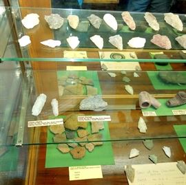 Lot 276: Large NC Cherokee Nation et al Relics and Arrowheads in Glass Case.
Impressive labeled collection of Native American relics, beads, tools, pottery shards and arrowheads and projectile points all excavated in various counties in North Carolina. Collection shows the county where items were found including Indian beads, spearhead and chisels from Hertford County, NC home of the Meherrin tribe; Cherokee drills and chisel from Yancey Co; a Native American sign found in Graham Co; an axe head found in guilford Co; a skinning knife and Tomahawk head from Yancey Co; a paint rock and game rock found in Cherokee Co; a gorget neck ornament from Clay Co; a spear head from Yancey Co; an axe head found in Madison Co; a Tomahawk from Madison Co; 3 peace pipes from various counties (see labels in photo); a Cherokee seal embroiered patch; a 28" wooden carved and painted Totem Pole. Top side of case has a crack from front to back about midway.
