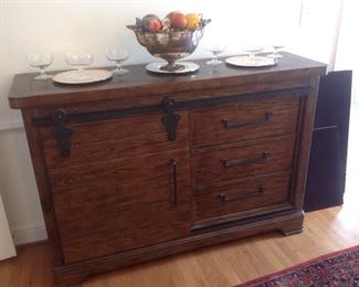 Dining Room:  This piece was brought up from the lower level to make it easier for the purchaser to remove.  The wooden cabinet has a barn-door on the left and three drawers on the right.  The barn door slides to the right to reveal three interior shelves.  This is a great storage piece!  It was formerly used to store electronics and was placed below a wall mounted TV but would also make an interesting kitchen peninsula. It measures 62" wide  18" deep x 42" tall.  The large stag horn silver-plate bowl filled with various balls (e.g., bocce, pool, bingo)is also for ale.  It is flanked by silver-plate chargers and a set of six large crystal dessert glasses.