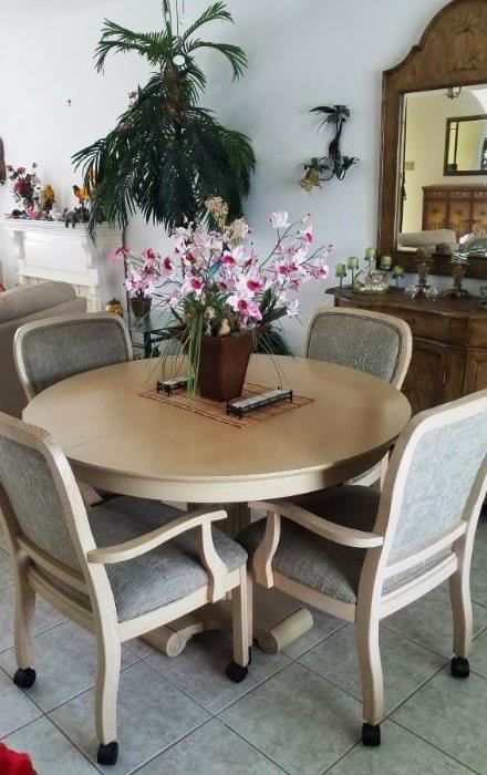 Round top light wood dining set with 6 rolling arm chairs(not sure if there are extra leaves yet).  Artificial pink orchid in brown pot. 