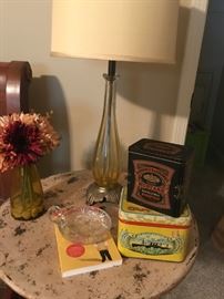 Small side table, table lamp, glass "nappy," and advertising tins.