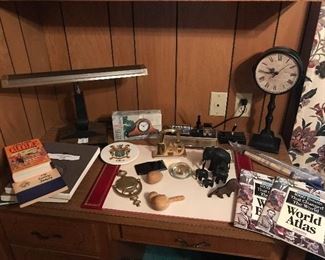 An assortment of desktop accessories which include an vintage ink well with pen and calendar display, a vintage calligraphy pen, a couple of clocks, lamp, Dad with horseshoe design letter holder, a couple of glass paperweights, a genealogy book to record family history, “World Atlas” and “World Events” books.