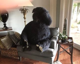 A large stuffed gorilla is comfortably seated in an upholstered armchair flanked by pillows, end table and a partially obscured fabulous floor lamp.  