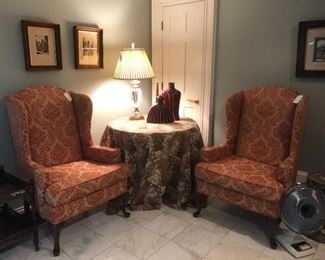 Medallion upholstered wing chairs fill a cozy corner along with a tapestry clothed table, acrylic table lamp, signed European street scene art, and metal trimmed end table.  