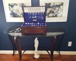 Glass top iron base demilune table displaying set of silverplate flatware. Spring flower artworks on wall.