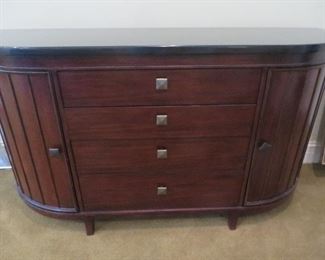 A Better Homes and Gardens shaped buffet with a dark charcoal acrylic top. Produced under license by Universal Furniture, this wood buffet features four center drawers with dovetail construction, and two curved end doors with vertical stiles. 69" W x 37.5" H x 20.75" D