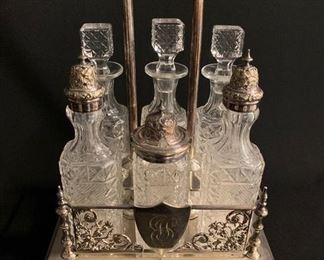 HIGH VICTORIA SILVER PLATE AND CUT GLAS CRUET STAND, Circa 1860.  The stand detailed with open work spandrels of scrolling tendrils and acanthus; two shields engraved with early monogram cyphers; the six original cut glass cruets with nicely chased silver caps and cover