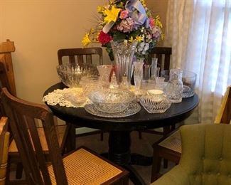 Black pedestal table w/ 5 antique wooden cane bottom chairs. On top of the table is an assortment of various types of crystal & glassware. Large silk flower arrangement in the middle of the table. Crocheted doily & yellow table runners. Green upholstered chair in lower right corner of picture. 