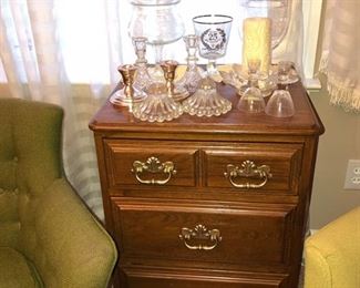 Bedside table with 3 drawers. Tall crystal dish w/ lid, candle sticks & flower shaped candle holder, glassware & pair of silver candle sticks.