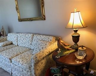 Light colored floral couch w/ 2 throw pillows, decorative antique gold mirror, 2 matching oval end tables, small lamp w/ shade, 2 matching bird figurines, small crystal bowl & antique wooden press.