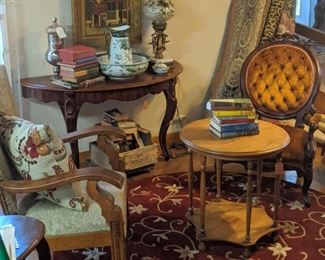 Large mahogany demi-lune entry table with two legs, round carved Center table, oak art Deco chair on the left and mahogany Victorian tufted back chair on the right. Machine-made round thick rug with floral design. ( Background numerous machine-made rugs) antique books and various lamps and vases.