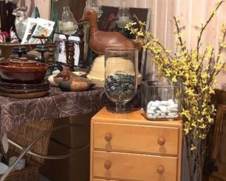 Small chest of drawers; cloth rooster; yellow flowers in a vase; clear glass decanters; stones
