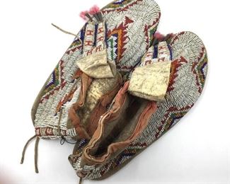 Pair of Native American Beaded Hide Moccasins, side sewn with rawhide sole, fully beaded with a geometric motif in red, orange, blue and green on white, with split tongues ended with tin cones. With cotton lining at ankle opening.  11 in. l.  Condition: In good condition slightly brittle, with some wear and deterioration, wear and deterioration to cotton lining at ankle.