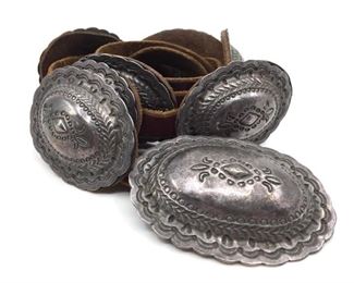 Silver Concho Belt, Of Futures Past. Silver Concho Belt, Of Futures Past, 1989, with stamped oval buckle and 9 similar oval leather backed concho plates, on thin brown leather strap, the buckle signed and dated, incised 999 ingot, with applied bear claw tag, each concho hook with bear claw tag.  Buckle is 3.5 x 2.75, each concho plate is 3 x 2 in. leather strap is 50 in. total length.  Condition: Buckle with bend at one end.