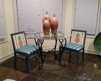 Pair of blue chairs with pineapple art on backs and glass top wrought iron table