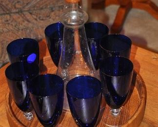 Round teak serving tray shown with a mid century glass and teak decanter and 8 cobalt blue beverage glasses 