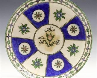 A Persian Ceramic Plate. Polychrome decoration with a paneled design and floral and foliate detail, in Blue, Green and Pink on a White ground, inscription at center. Wear and crazing with some staining, hairlines and an old repair to rim, rim flakes and one 1/2" chip. 9 3/4" diameter.