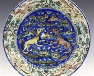 A Persian Ceramic Plate. Colorful plate is decorated in azure, yellow, pinkish mauve, and green and contains images of Jackals amidst plants and trees at the well which is contained within a border of similar fashion. Crazing, two chips to the rim. 8 1/2" diameter.