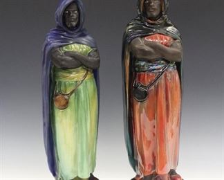Two Royal Doulton Porcelain Figures of Moors. Both figures of men, in the same pose, but of slightly different sizes; the small figure is by Charles Noke for Royal Doulton, and wears an electric blue cloak with a green belt and garment, the larger figure wears a multicolor cloak, consisting of black, red, green, blue, and yellow, a multicolor belt and reddish orange garment, with tinges of green and yellow. Both with Royal Doulton trademark stamp, the first also with "HN 33 'An Arab'-C. J. Noke. & C. Potted by:-Doulton & Co." and the second with "The Moor" in gold at the underside. Very minor wear overall. 16 & 17" high.