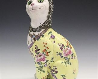 A Late 19th Century Galle Faience Cat. Figure of a seated cat, with glass eyes, is painted with ornate floral and geometric decoration at the back of the head, a yellow body with various types of floral sprays in multiple colors, and a collar with a locket suspended from it containing a portrait of a dog. Crazing overall, light wear to the decoration. 13" high.