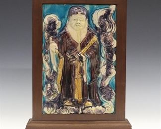 A Chinese Glazed Ceramic Tile. Rectangular molded tile with Daoist Immortal in high relief shown among the clouds, holding a fly whisk in his right hand and a scroll in his left, with Fahua inspired glaze overall, in colors of eggplant, yellow, and turquoise. Unmarked. Damaged and repaired at the lower right corner. Tile is 7 3/4 x 11" high, set within a modern wood frame with some damage, 10 x 12 3/4" high overall.