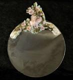 ROUND WALL MIRROR. GERMAN PORCELAIN FIGURE OF A LADY AND MULTI COLORED FLOWERS DECORATE THE TOP OF THE MIRROR. GOOD. 20" H X 16" W.
