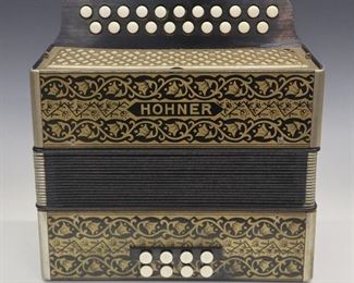 Lot 101: An early 20th century Hohner accordion.  21 key, 2 row, 8 Bellow model with Gold "Pokerwork" painted decoration. Minor surface wear, leather pull strap and one closure strap are present but broken.  11" high