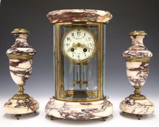 A turn of the century French three piece clock set with a crystal regulator and two garnitures.  8-day time and strike movement with a painted flat porcelain dial and Arabic numerals, fancy gilded hands and faux mercury pendulum.  Oval brass case with beveled glass panels, molded Marble top and base, garnitures with turned marble bodies and bronze fittings.  Minor wear, running when cataloged.  12 3/4" high overall.  ESTIMATE $600-800