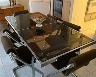 Vintage dining table in the style of Milo Baughman constructed of smoked chrome-plated steel with extendable top of three black glass panes trimmed in brass. Two panes easily slide horizontally revealing a hidden middle pane which pivots into place. Four quarter-round smoked chrome legs. Robust, steady, sturdy. Two end leaves show signs of heavy use and scratches to gunmetal frame as well as black glass. Middle leaf in overall very clean condition from apparent lack of regular use. Table is 66" long by 40" wide. 98" long including 32" leaf. $2000