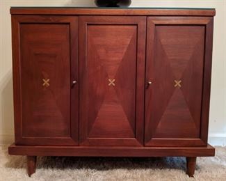 Close-up of the American of Martinsville two door walnut chest, showing inlaid brass "x" detail.                    The clean lines and beautiful hardwoods of this furniture makes American of Martinsville quite desirable today. Typically, you will see the company used oak as their secondary wood (inside the drawers) and gave preferential treatment to walnut as the primary wood.
You may notice that quite a few American of Martinsville pieces have something in common. That is their famous brass inset “x’s.” Often these “x’s” were inlaid in table tops, dresser corners and even on credenzas.