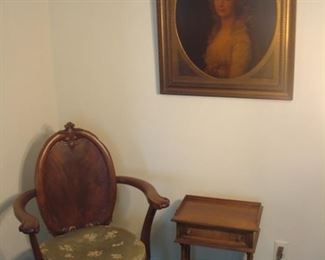 Living Room:  An antique wooden chair with detached  seat cushion is next to a vintage table with one drawer and magazine rack.  Above is a reproduction "painting" on board.  