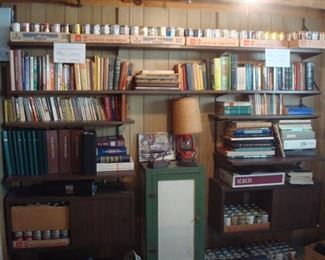 Lower Level:  Flats of empty beer cans are on the top shelf and on the floor, framing lots of books.  Also shown are two sad irons; a lantern lamp; and a green wooden cabinet.