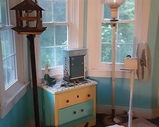 Decorative bird cage with stand. Small dresser with mosaic top. Knick knacks and other decorations. Tall lamp. Oscillating fan. Bed.