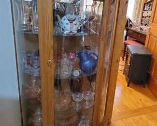 Antique curved glass china cabinet, items inside are individually priced. Pink depression glass, various pieces of glass and dishes. On top of cabinet is a basket of multi-colored marble eggs.