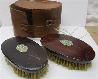 A pair of Deco clothing brushes with leather case, each with Art Nouveau style stamped metal ornaments.  Some bristles missing, wear to finish from use