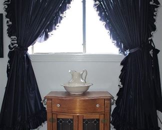 Pulaski Furniture Company Keepsake Collection Golden Oak Wash Stand with Brass Rod Towel Bars on each end,  Leaded Stain Glass Double Doors (30”W x 20”D x 28”H) shown with an Opalescent Glazed Ceramic Large Pitcher and Bowl Set.  Dorothy Ruffled Originals Navy Priscilla Style Curtains