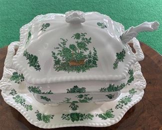 This tureen is an oblong octagonal shape with a handled lid, floral embossed handles, and four feet on the base. The body and lid of the tureen are decorated with a design depicting a basket of green flowers and plants. This tureen comes with a matching underplate and both pieces are marked “Spode Copeland China England”. The underplate measures 15" x 11.5" 