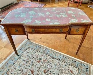 $475 -Louis XVI style writing desk with 3 drawers, brass ring pulls; fabric panel under safety glass; band inlay, raised on brass capped feet and casters; 30" H x 49" W x 23.5" D