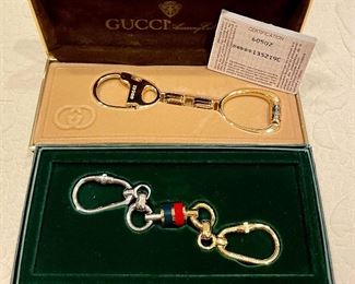 Item 483:  (2) Gucci Key Ring: $75 each                                    a. (bottom) Gold and Silver Tone                                                     b. (top) Gold against Tan Background