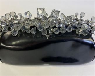 Lot	217	Prada Black Vernice Crystal Kiss Lock Clutch	Vernice black patent leather Prada clutch with silver-tone hardware, detachable flat leather strap with buckle adjustment, crystal embellishments at frame, blush leather lining, dual interior pockets; one with zip closure and crystal kiss-lock closure at top. One of the crystal embellishments is detached. Some minor scratches and marks. 12" l x 7" h x 4" w Some specks and indents on leather and appears to be glue marks on all top 4 seams on closure
