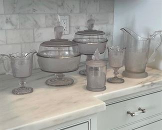 (6pc) STUNNING "TALLY-HO" PRESSED GLASS | pressed with scenes of log cabins, deer and bison, and with kneeling native american figure finials; including two lidded compotes (tallest 12-1/2 in., over finial) plus a pitcher (with small chip to foot), a small pitcher, goblet, and lidded beaker 