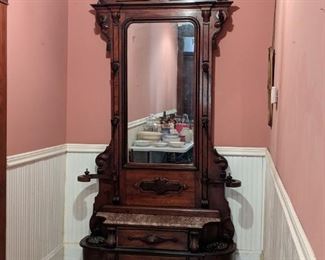 Hand-carved Walnut hall tree, beveled mirror, marble top seating, and original iron drip pans.